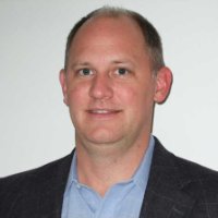 A man with short light brown hair wearing a dark jacket and light blue collared shirt, standing in front of a plain white background.