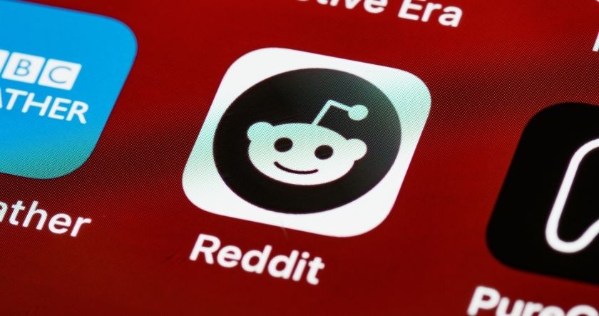Close-up of the Reddit app icon displayed on a mobile device screen, with a red background and other app icons partially visible.