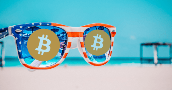 A pair of sunglasses with an American flag design has Bitcoin symbols over the lenses, with a blurred beach and ocean in the background.