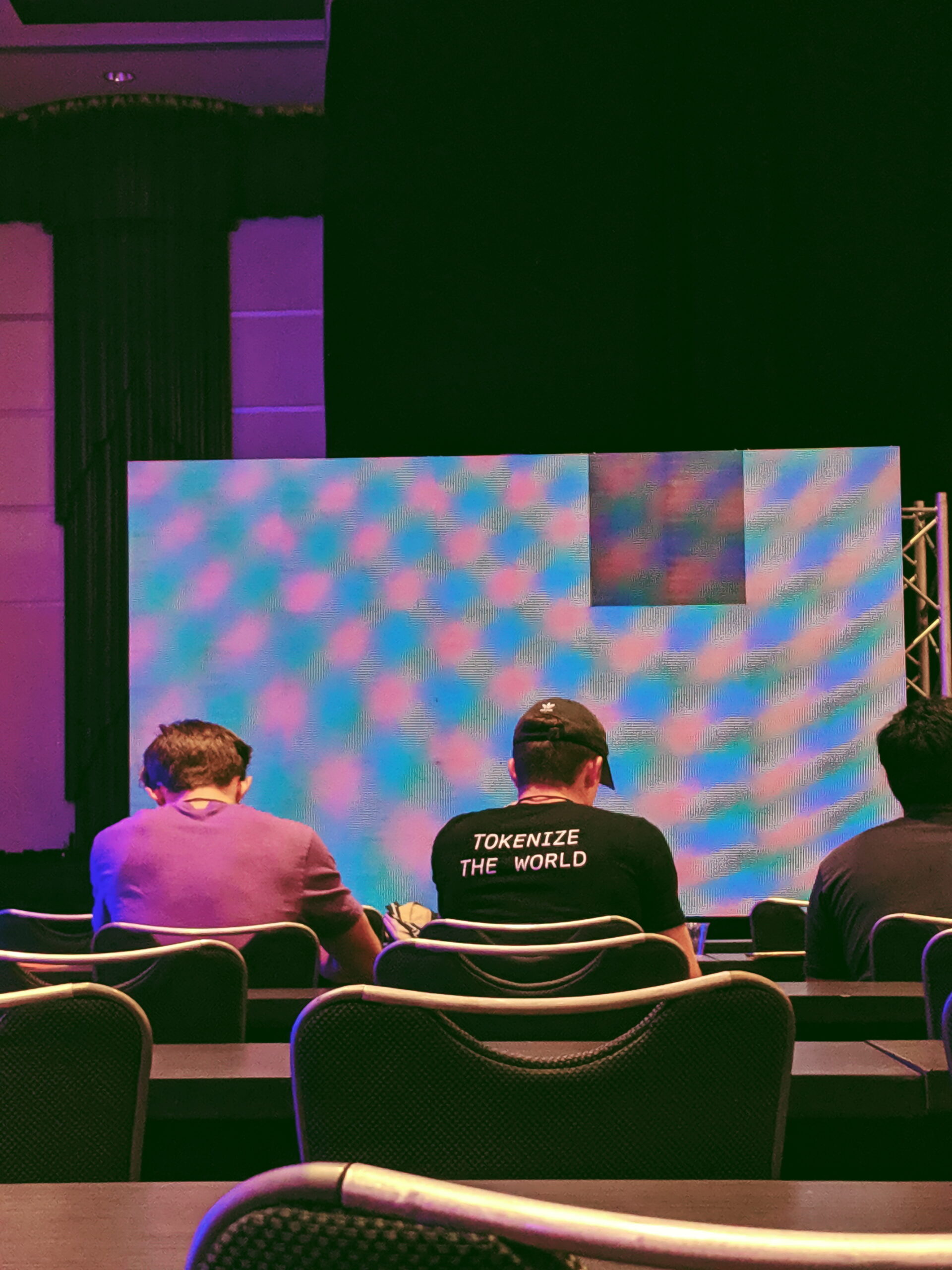 Three people sit in front of an empty stage with a large screen displaying a blue and purple pattern. The person in the center wears a shirt that says "TOKENIZE THE WORLD.