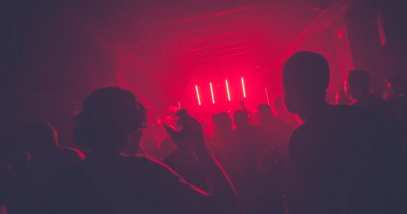 A crowd of people stands and dances in a dark room with red stage lights illuminating the space.