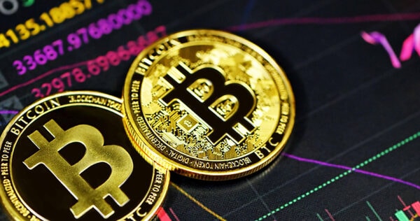 Two gold-colored Bitcoin tokens placed on a surface with colorful cryptocurrency price charts and numbers in the background.