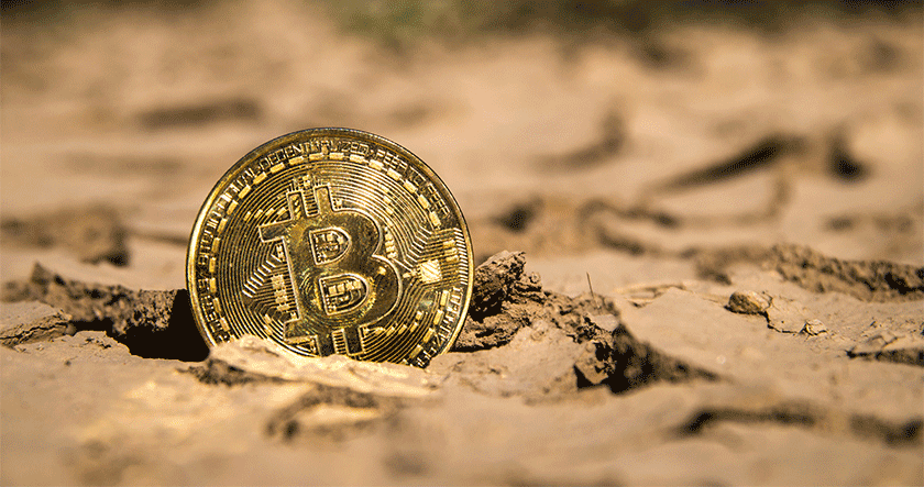 A gold Bitcoin coin partially embedded in dry, cracked earth.