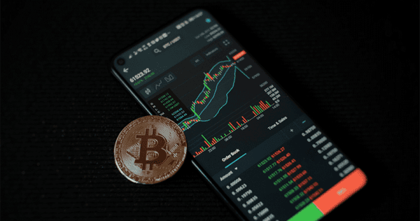 A smartphone displays a cryptocurrency trading chart and data, with a physical Bitcoin coin placed next to it on a dark surface.