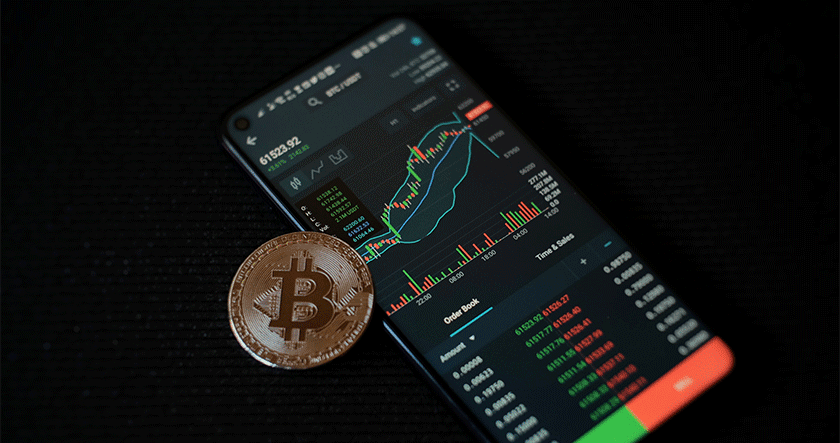 A smartphone displays a cryptocurrency trading chart and data, with a physical Bitcoin coin placed next to it on a dark surface.