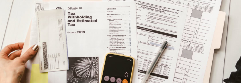 A person holds a check next to tax forms, a publication titled "Tax Withholding and Estimated Tax," a pen, and a smartphone with a calculator app open.