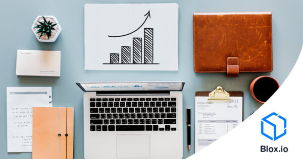 Top view of a desk with a laptop, documents, notebook, pen, coffee cup, plant, business cards, leather folder, and a chart showing upward growth, with a Blox.io logo in the corner.