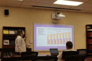 A woman stands in front of a projected presentation displaying a bar chart, speaking to a seated man with a laptop in a classroom or conference room setting.