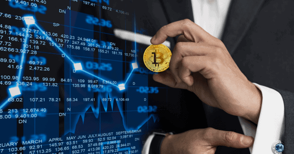 A person in a suit holds a Bitcoin coin in front of a digital screen displaying financial charts and stock market data.