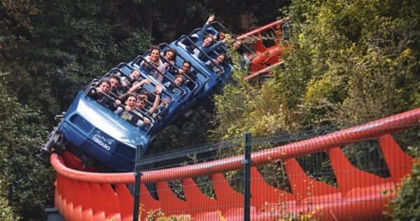 A blue roller coaster train filled with people rounds a curved, red track surrounded by greenery; several riders have their hands raised.