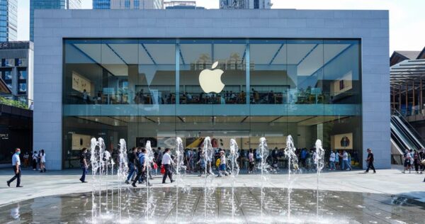Street-level view of an Apple Store with a large glass facade, the Apple logo above the entrance, people walking by, and water fountains in the foreground.