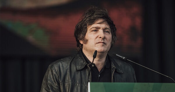 A man with medium-length brown hair, wearing a black leather jacket, stands at a podium with microphones and speaks in front of a blurred background.