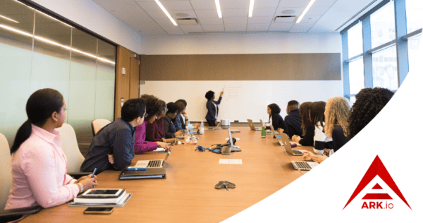 A group of people sit around a conference table with laptops, watching a presenter write on a whiteboard; ARK.io logo is visible in the bottom right corner.