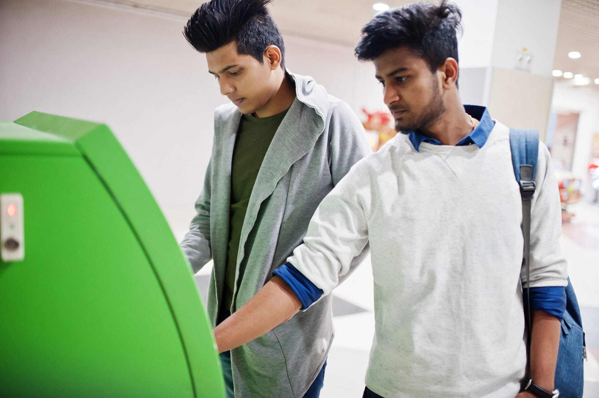 Two young men stand at a green ATM or self-service kiosk. One man is using the machine while the other stands beside him, watching and waiting. They appear to be indoors in a public or commercial space.