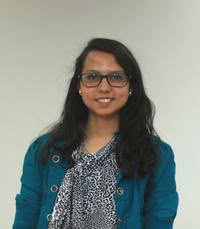 A woman with long dark hair and glasses, wearing a patterned blouse and teal jacket, stands in front of a plain light-colored background.
