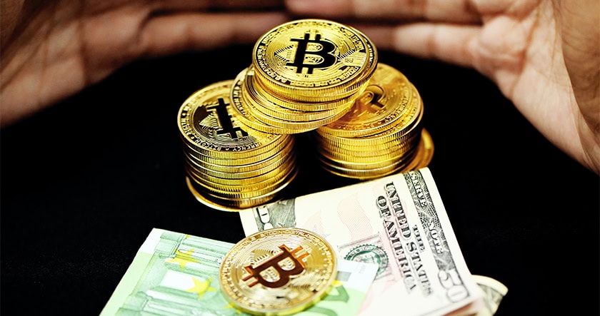 Stacks of gold-colored Bitcoin tokens are placed on top of U.S. dollar bills with hands partially visible in the background.