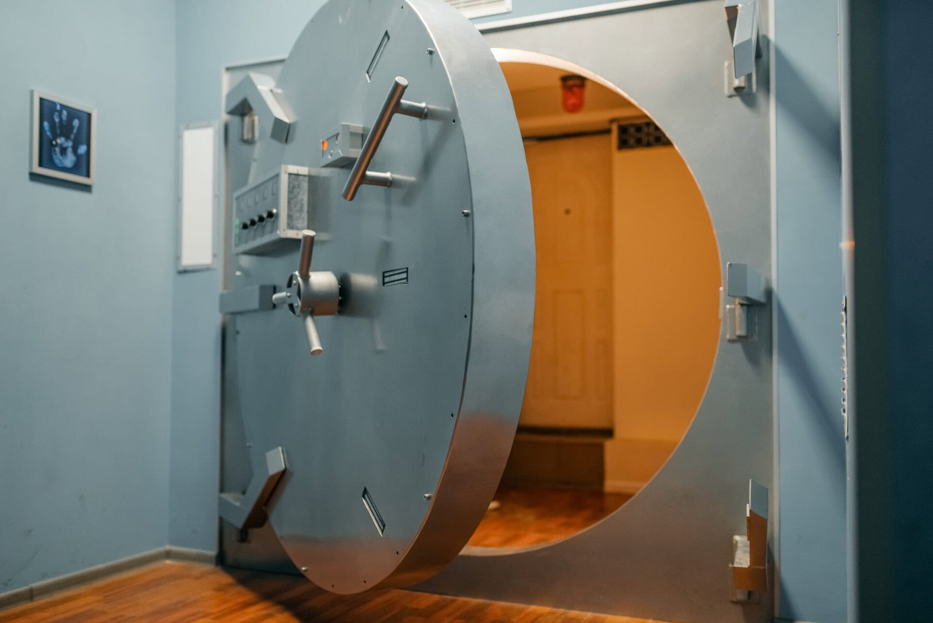 A large, round metal vault door stands open, revealing a secure room with wooden floors and beige walls. The surroundings are painted blue, and a framed picture is visible on the wall to the left of the door.