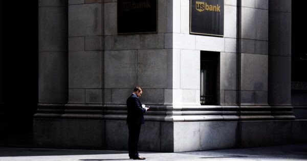 A man in a suit stands alone on a city sidewalk in front of a large stone building with two US Bank signs, looking at his phone.