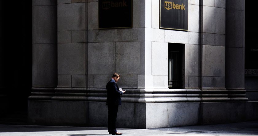 A man in a suit stands alone on a city sidewalk in front of a large stone building with two US Bank signs, looking at his phone.