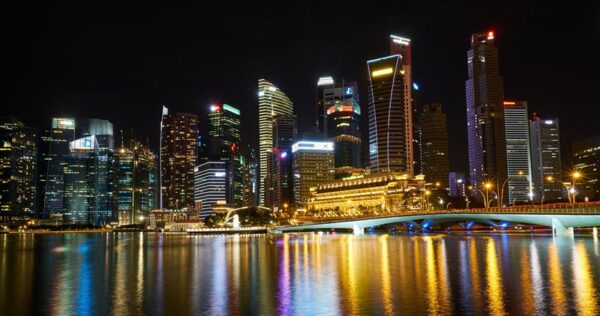 Night view of a city skyline with illuminated skyscrapers and their reflections on the water, featuring a bridge in the foreground.