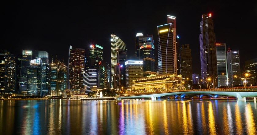 Night view of a city skyline with illuminated skyscrapers and their reflections on the water, featuring a bridge in the foreground.