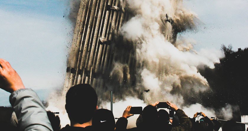 A large building is being demolished, collapsing amid clouds of dust, while a crowd of people stands in the foreground taking photos and videos.