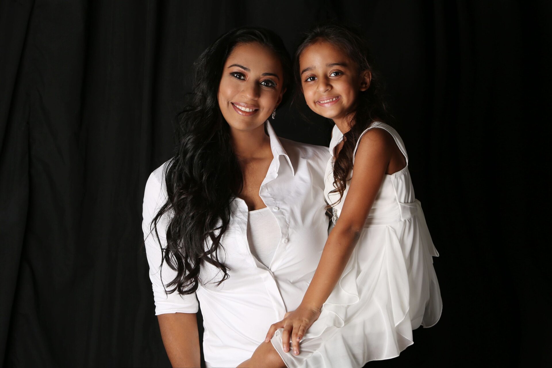 A woman in a white shirt and a young girl in a white dress smile together in front of a black background.