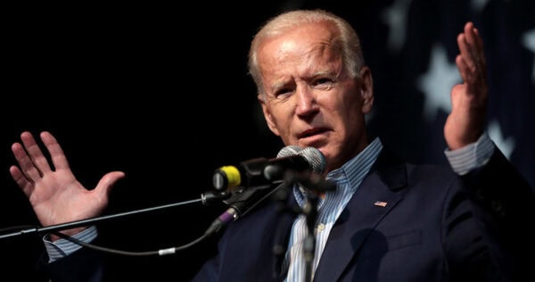 A man in a suit speaks into a microphone with his hands raised, standing in front of a dark background with white stars.