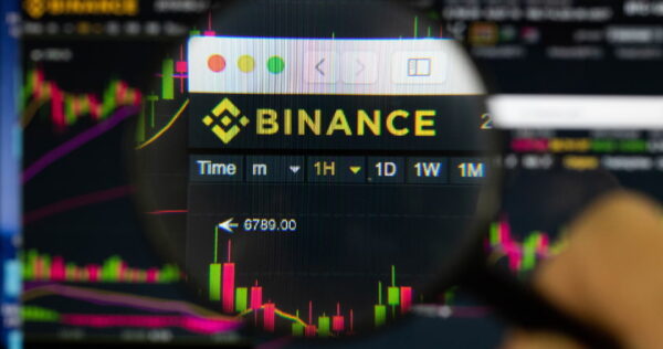 A magnifying glass focuses on the Binance cryptocurrency trading platform interface, showing price charts and trading options on a computer screen.
