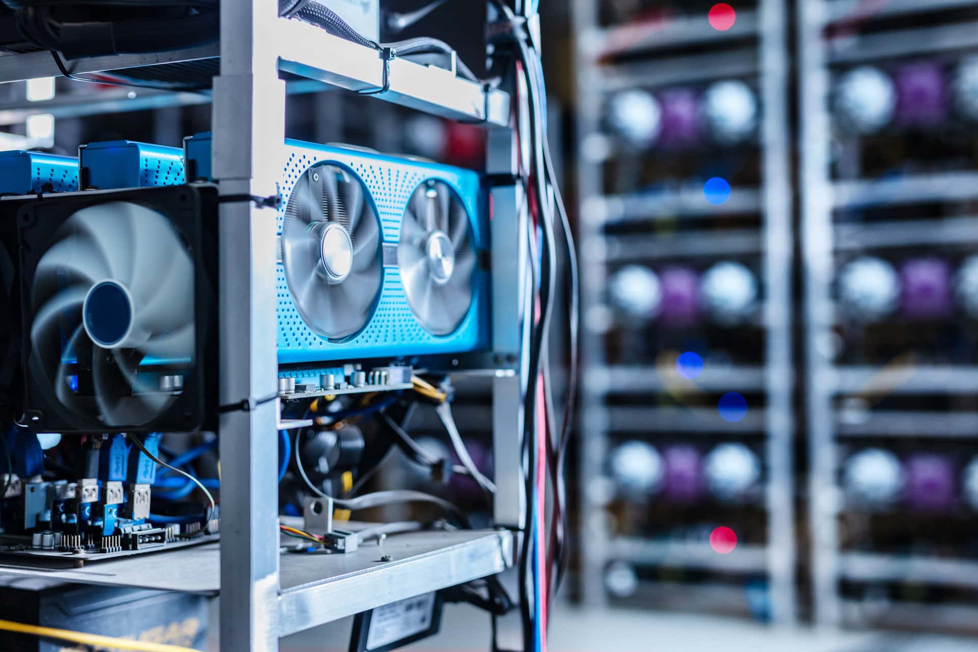 Close-up of computer graphics cards and fans on a metal rack, with multiple similar racks blurred in the background, likely inside a cryptocurrency mining facility.