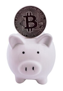 A white piggy bank with a large Bitcoin coin inserted in the slot, symbolizing cryptocurrency savings or investment. The background is white.