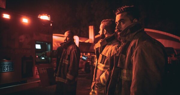 Three firefighters in protective gear stand near an emergency vehicle at night, illuminated by red lights.