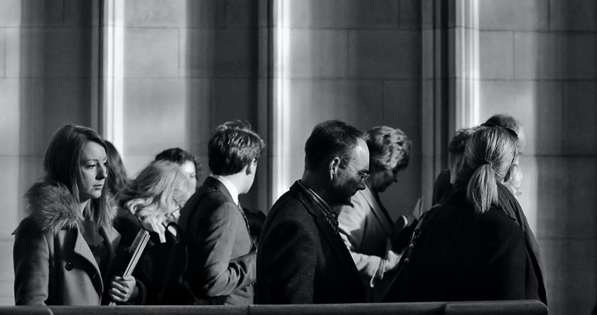 A group of people in coats, some holding notebooks, stand and talk in a sunlit corridor with tall stone columns.