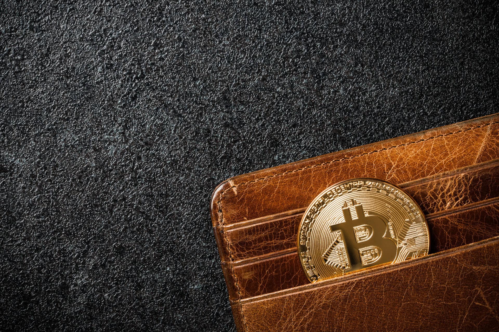 A shiny gold Bitcoin coin partially tucked into a brown leather wallet, set against a dark textured background.