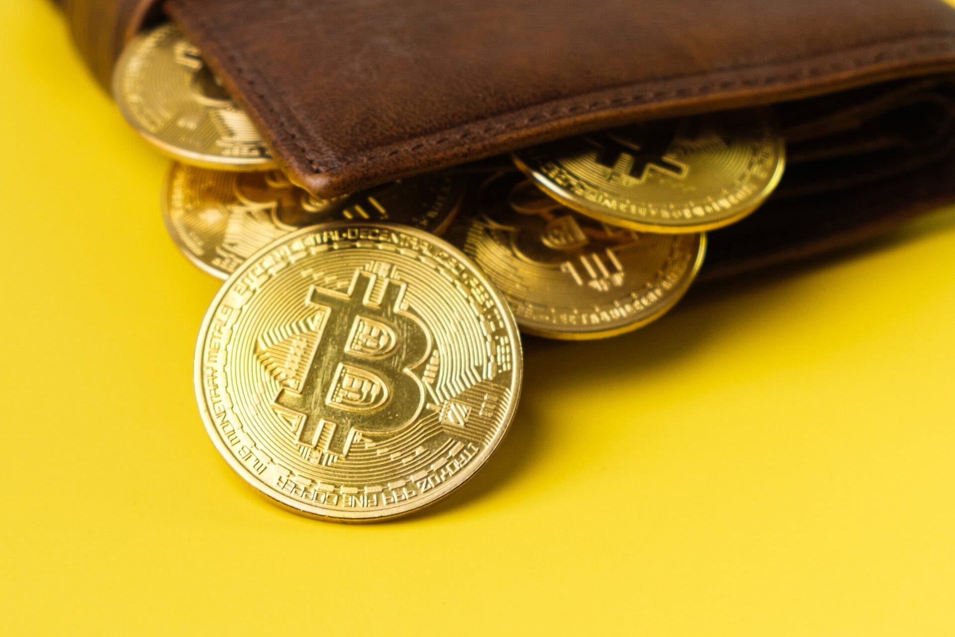 A brown leather wallet with several gold-colored Bitcoin coins spilling out on a bright yellow background.