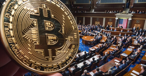 A large physical Bitcoin coin is held in focus in front of a blurred scene of a legislative assembly with many seated officials and the U.S. flag visible.