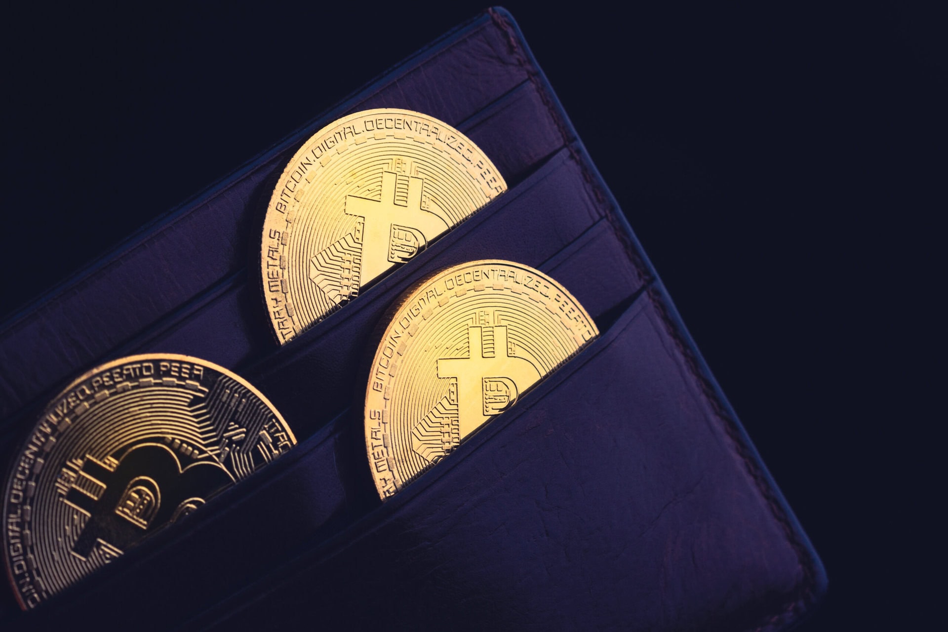 Close-up of a dark leather wallet with three gold-colored Bitcoin tokens partially visible in its card slots, symbolizing digital currency and cryptocurrency storage.