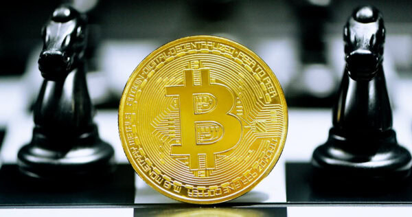 A gold Bitcoin coin stands upright on a chessboard between two black knight chess pieces.
