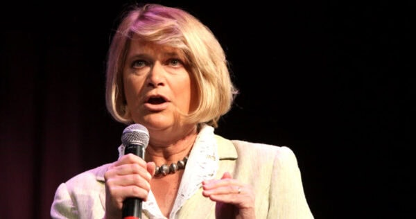 A woman with blonde hair holds a microphone and speaks on stage, wearing a light-colored blazer and necklace against a dark background.