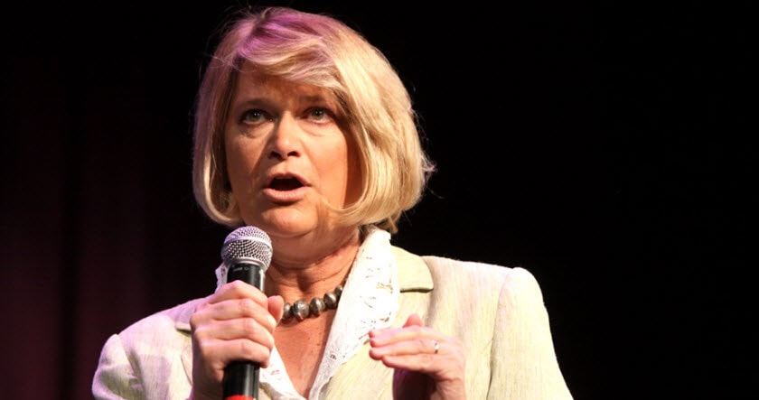 A woman with blonde hair holds a microphone and speaks on stage, wearing a light-colored blazer and necklace against a dark background.