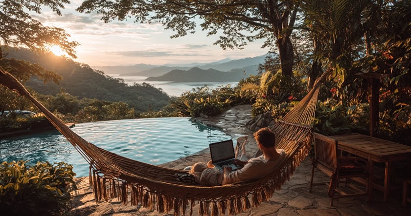A person relaxes in a hammock with a laptop by an outdoor pool, surrounded by lush greenery and overlooking a scenic mountain and lake view at sunset.