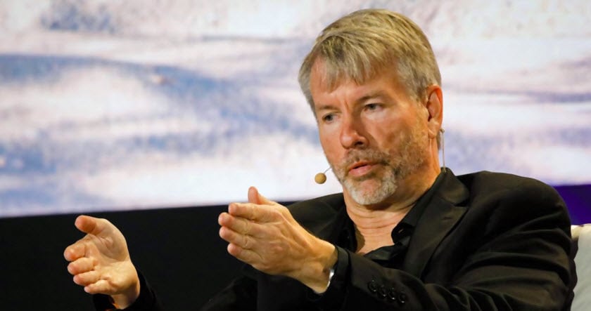 A man with a headset microphone, wearing a black jacket, gestures with his hands while speaking on stage.