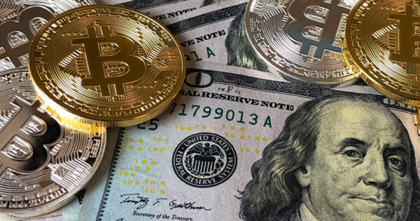 Several gold-colored physical Bitcoin tokens rest on top of US one hundred dollar bills featuring Benjamin Franklin's portrait.