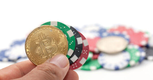 A hand holding a gold Bitcoin coin and several colorful poker chips, with more chips blurred in the background.