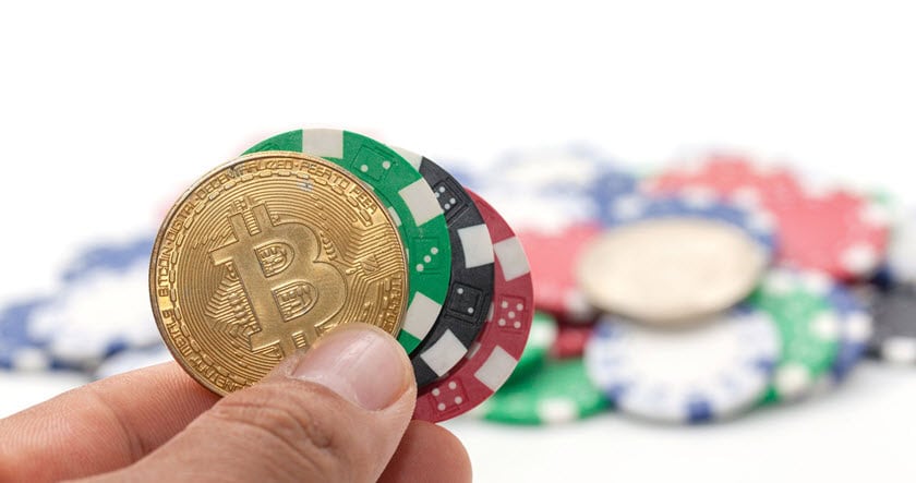 A hand holding a gold Bitcoin coin and several colorful poker chips, with more chips blurred in the background.
