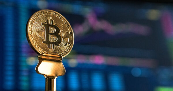 A gold-colored Bitcoin coin is displayed in the foreground with a blurred digital stock chart in the background.
