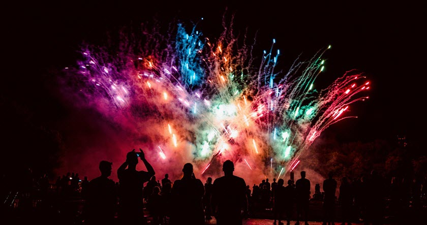 A crowd watches colorful fireworks lighting up the night sky, with vibrant bursts of red, green, blue, and purple visible above silhouetted spectators.