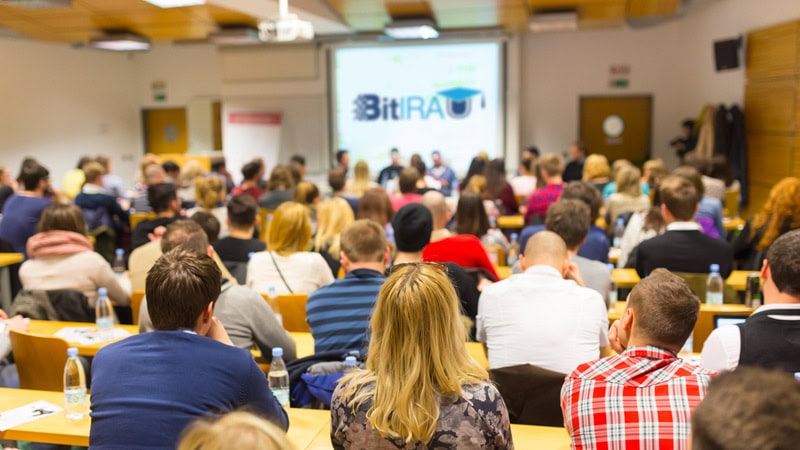 A group of people sit in rows facing a panel at the front of a classroom or seminar room, with a presentation projected on a screen.