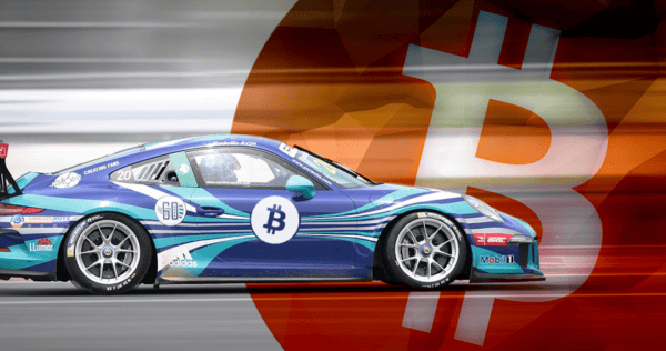 A blue race car with a Bitcoin logo drives past a large, blurred Bitcoin symbol in the background.