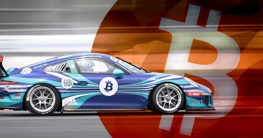A blue race car with a Bitcoin logo drives past a large, blurred Bitcoin symbol in the background.